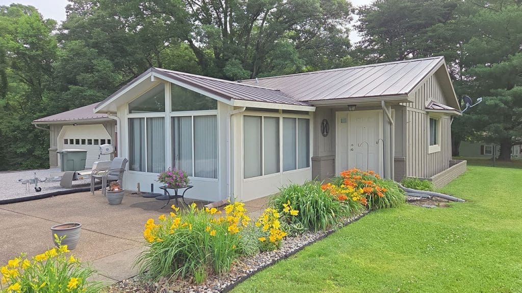 Front exterior repaint in Oakland City IN showing light sage green siding, white entry door, sunroom windows, and brown metal roof