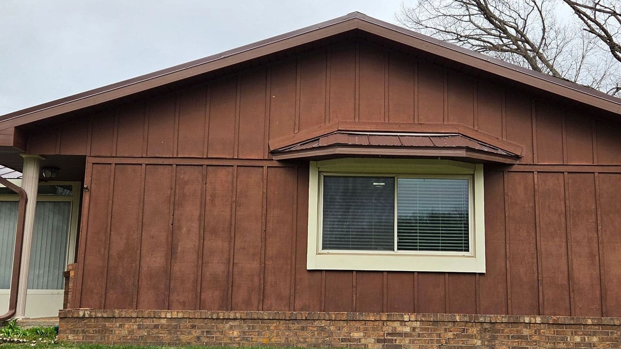 Before exterior house painting project showing faded brown vertical board and batten siding and brick foundation