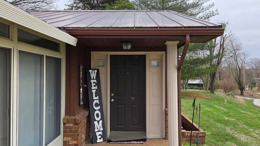 Before front door exterior repaint in Oakland City IN showing dark entry door, welcome sign, and brown porch trim under metal roof